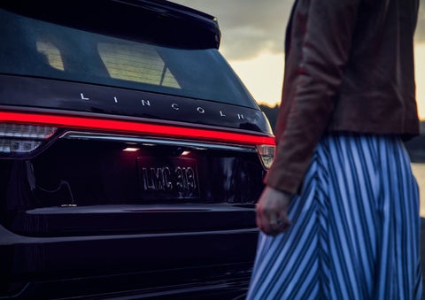 A person is shown near the rear of a 2024 Lincoln Aviator® SUV as the Lincoln Embrace illuminates the rear lights | Johnson Sewell Lincoln in Marble Falls TX