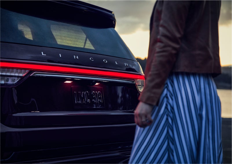 A person is shown near the rear of a 2023 Lincoln Aviator® SUV as the Lincoln Embrace illuminates the rear lights | Johnson Sewell Lincoln in Marble Falls TX